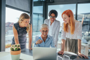Diverse group of professional women smiling and collaborating around a laptop in a bright modern office, symbolizing teamwork and employee engagement.